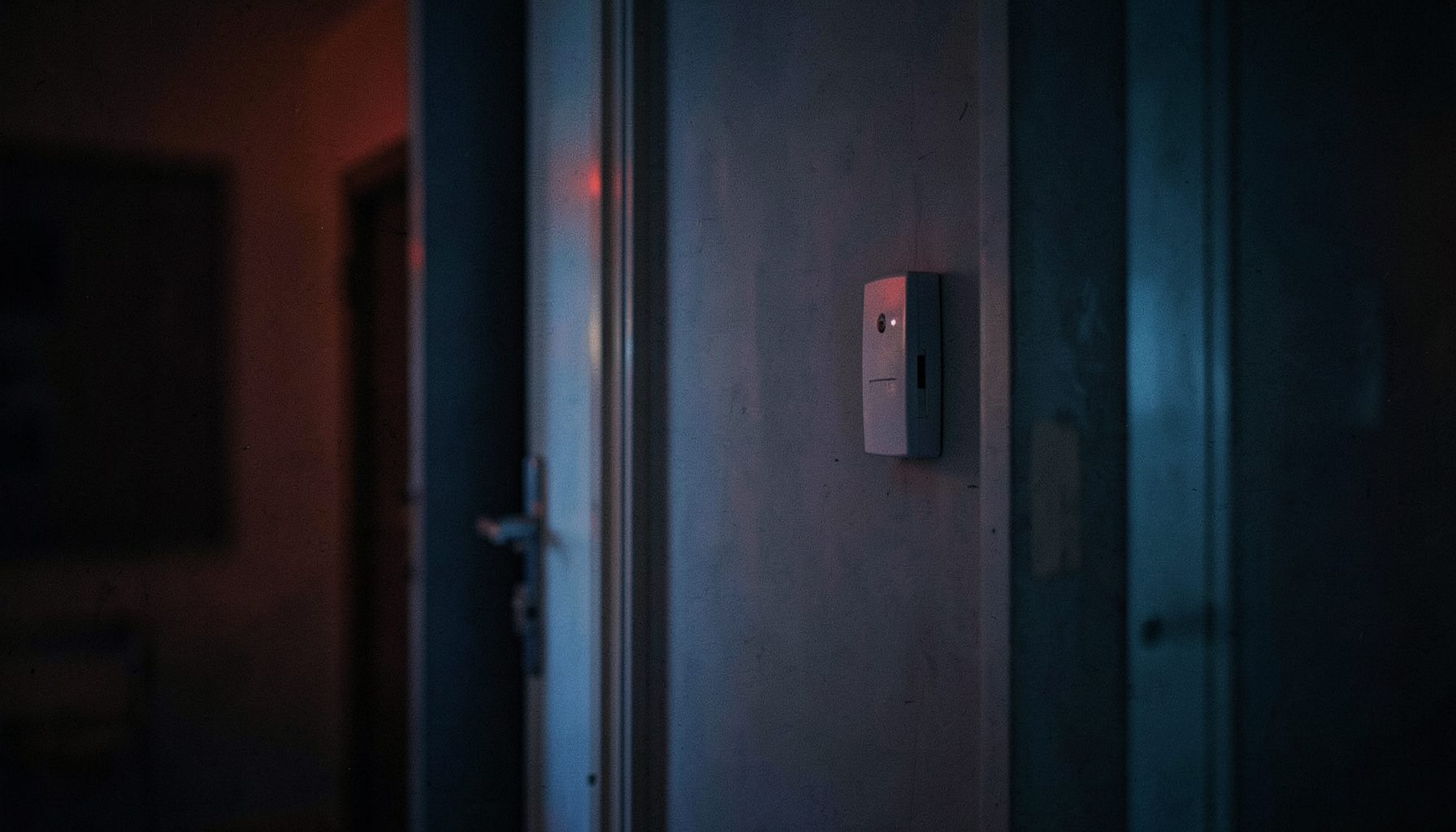 Intérieur résidentiel tamisé au crépuscule, un boîtier de centrale d'alarme discret fixé au mur près d'une porte, ambiance sécurisante aux tons bleu nuit et gris chauds.