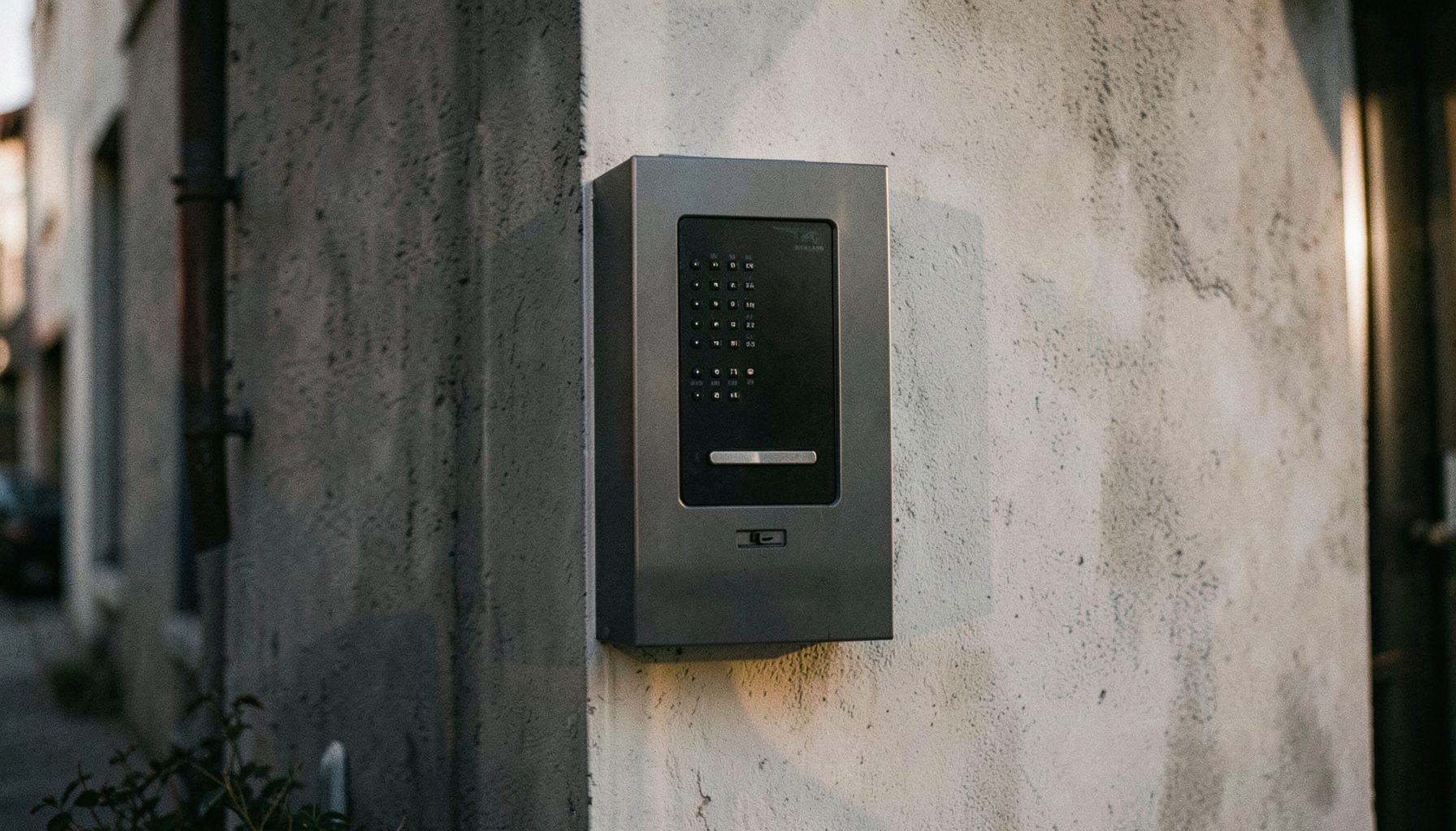 Platine de rue interphone en saillie sur un mur extérieur, tons gris anthracite et blanc cassé, lumière douce de fin d'après-midi