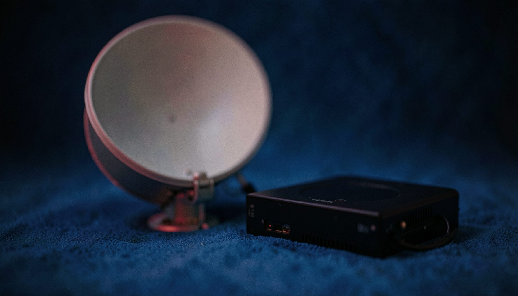 Gros plan doux sur une parabole satellite grise et un boîtier récepteur noir posés sur un fond bleu foncé feutré